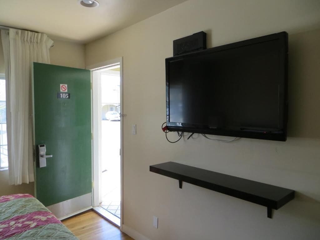 A room with a bed, a television mounted on the wall, a door with a number sign, and a window with curtains.