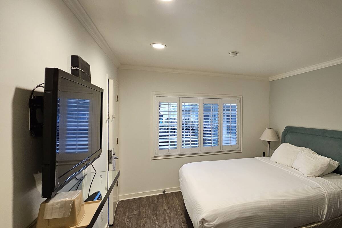 A hotel room with a bed, a television, a window with blinds, and a lamp.