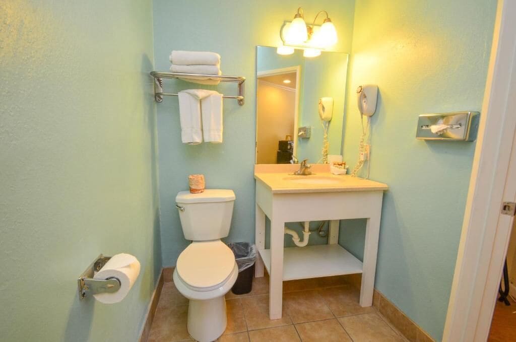 A bathroom with a toilet, sink, and mirror. There is a towel rack with towels, a trash can, and a toilet paper holder.