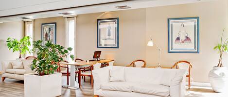 A spacious living area with a white sofa, a dining table, and chairs. There are framed artworks on the walls and a potted plant in the center.