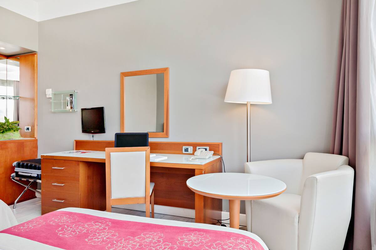A hotel room with a bed, a desk with a chair, a small table, a mirror, a lamp, and a TV.