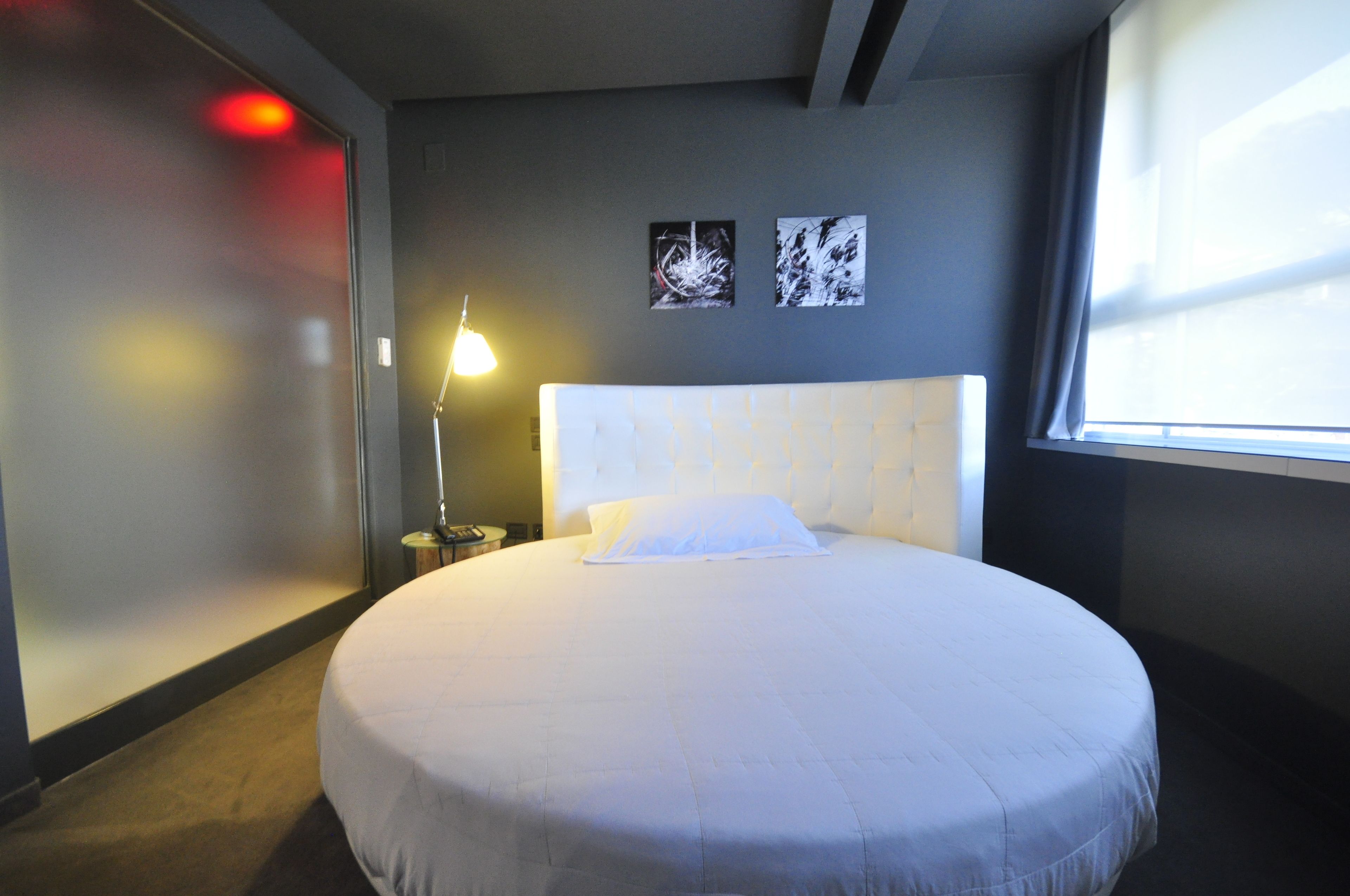 A hotel room with a round bed, a bedside lamp, and two abstract art pieces on the wall.