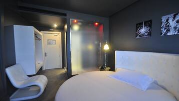A modern hotel room with a large bed, a white armchair, a bedside table, and a door leading to a bathroom.