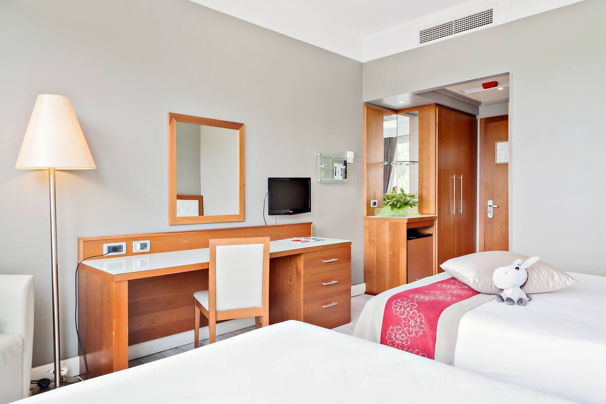 A hotel room with a wooden desk, a chair, a bed with a red and white pillow, a small stuffed animal, and a TV.