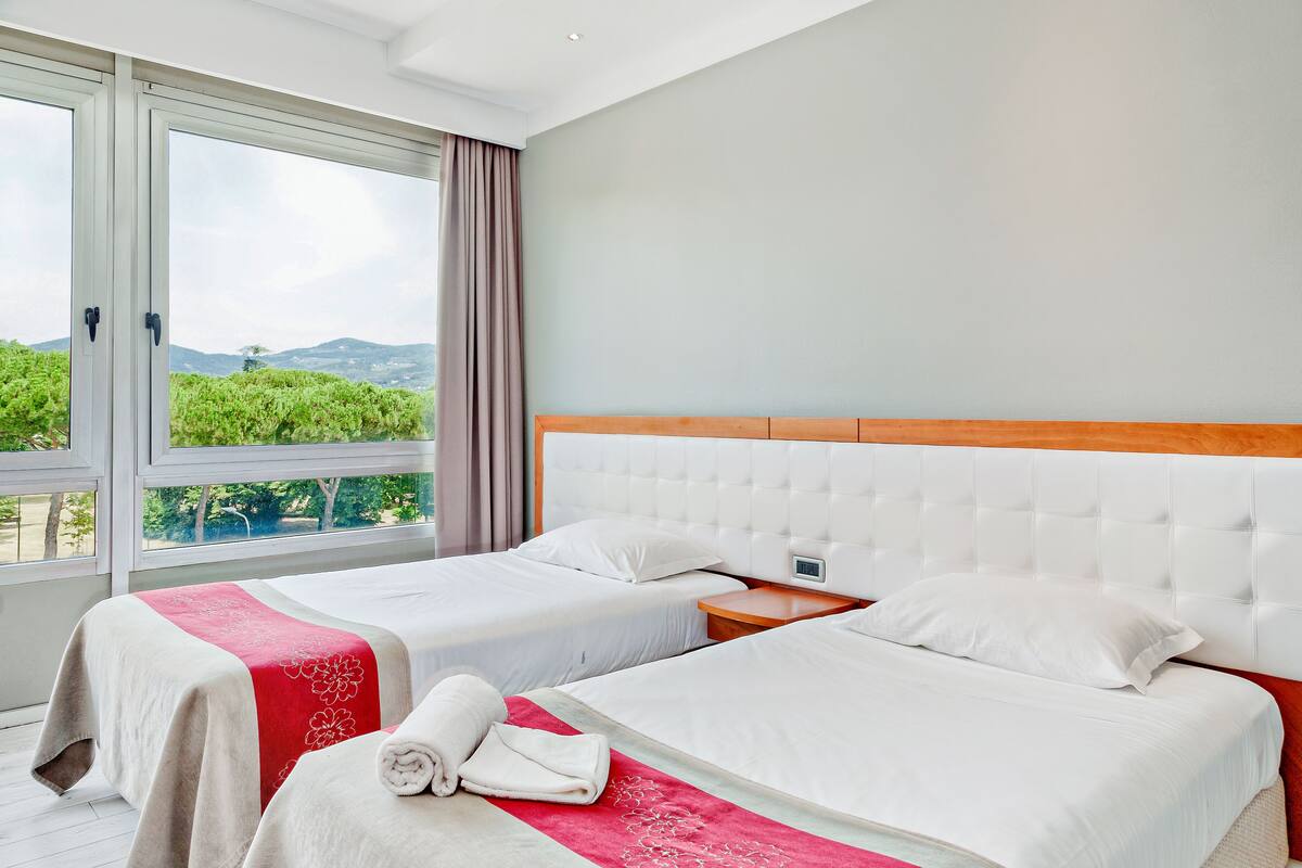 A hotel room with two beds, a large window with curtains, and a view of greenery and mountains.