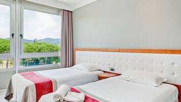 Ein Hotelzimmer mit zwei Betten, einem großen Fenster mit Vorhängen und Blick auf Grünflächen und Berge.