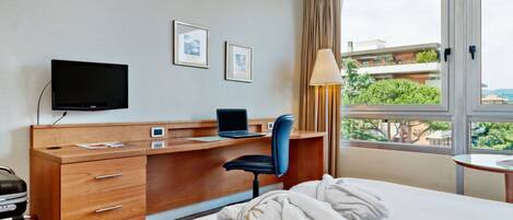 Habitación de hotel con cama, escritorio con laptop, televisión y ventana con vista a edificios y árboles.