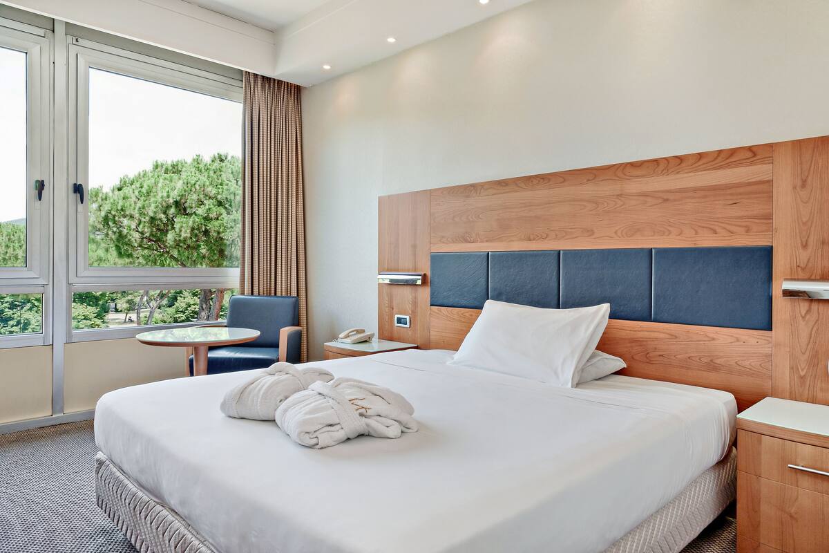 A hotel room with a large bed, a wooden headboard, a small table, a chair, and a view of trees through the window.