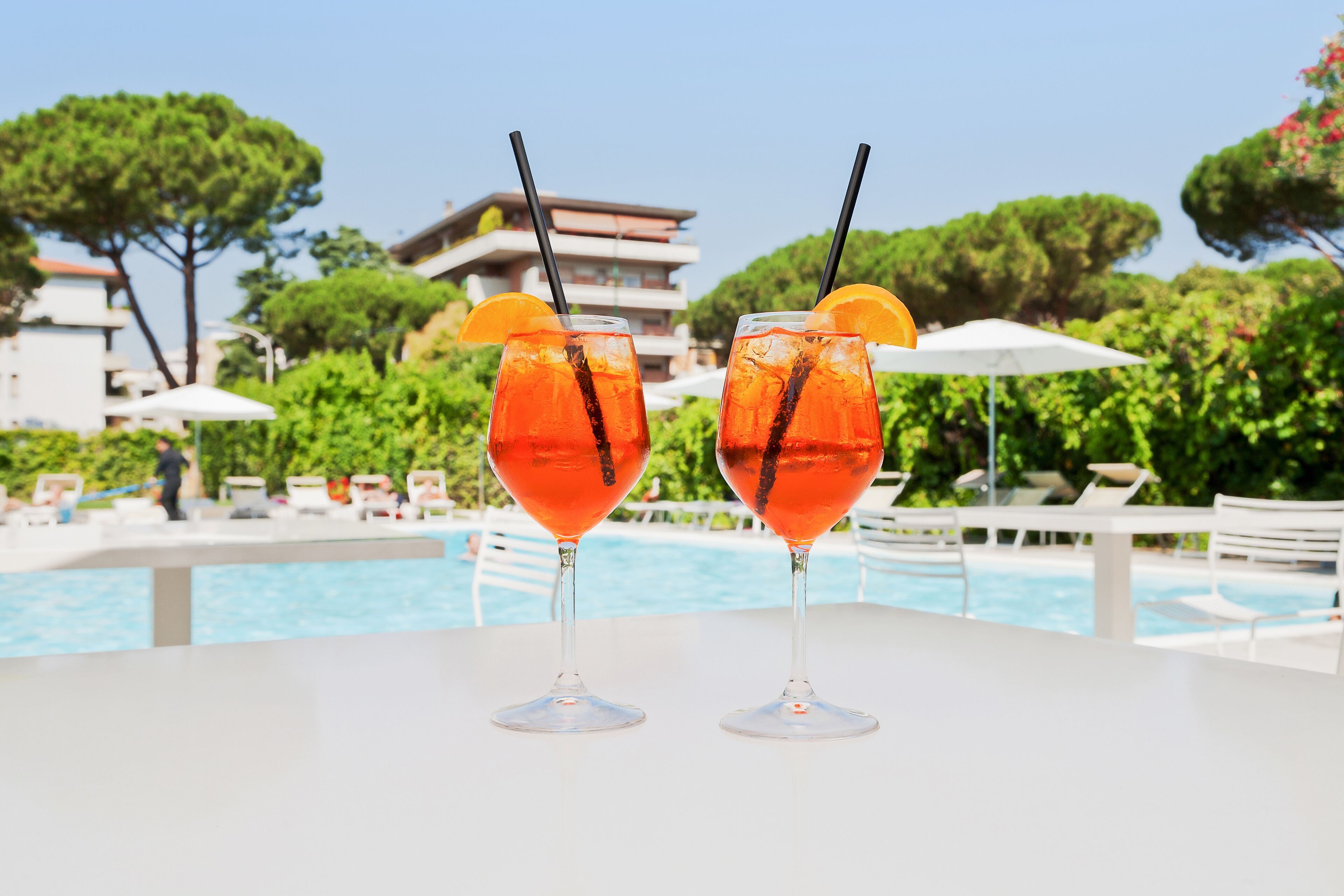 Dos vasos de bebida roja con pajitas y rodajas de naranja sobre una mesa blanca junto a una piscina con sillas de descanso y sombrillas.