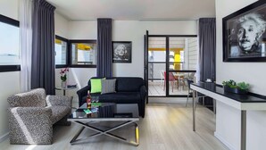 A modern living room with a black sofa, patterned armchair, and a glass coffee table. There is a dining area with a white table and chairs, and a balcony with yellow chairs. - Best Western Regency Suites (Tel Aviv)