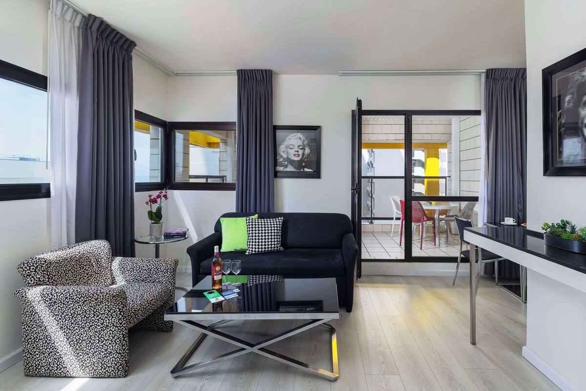 A modern living room with a black sofa, patterned armchair, and a glass coffee table. There is a dining area with a white table and chairs, and a balcony with yellow chairs.