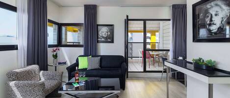 A modern living room with a black sofa, patterned armchair, and a glass coffee table. There is a dining area with a white table and chairs, and a balcony with yellow chairs.