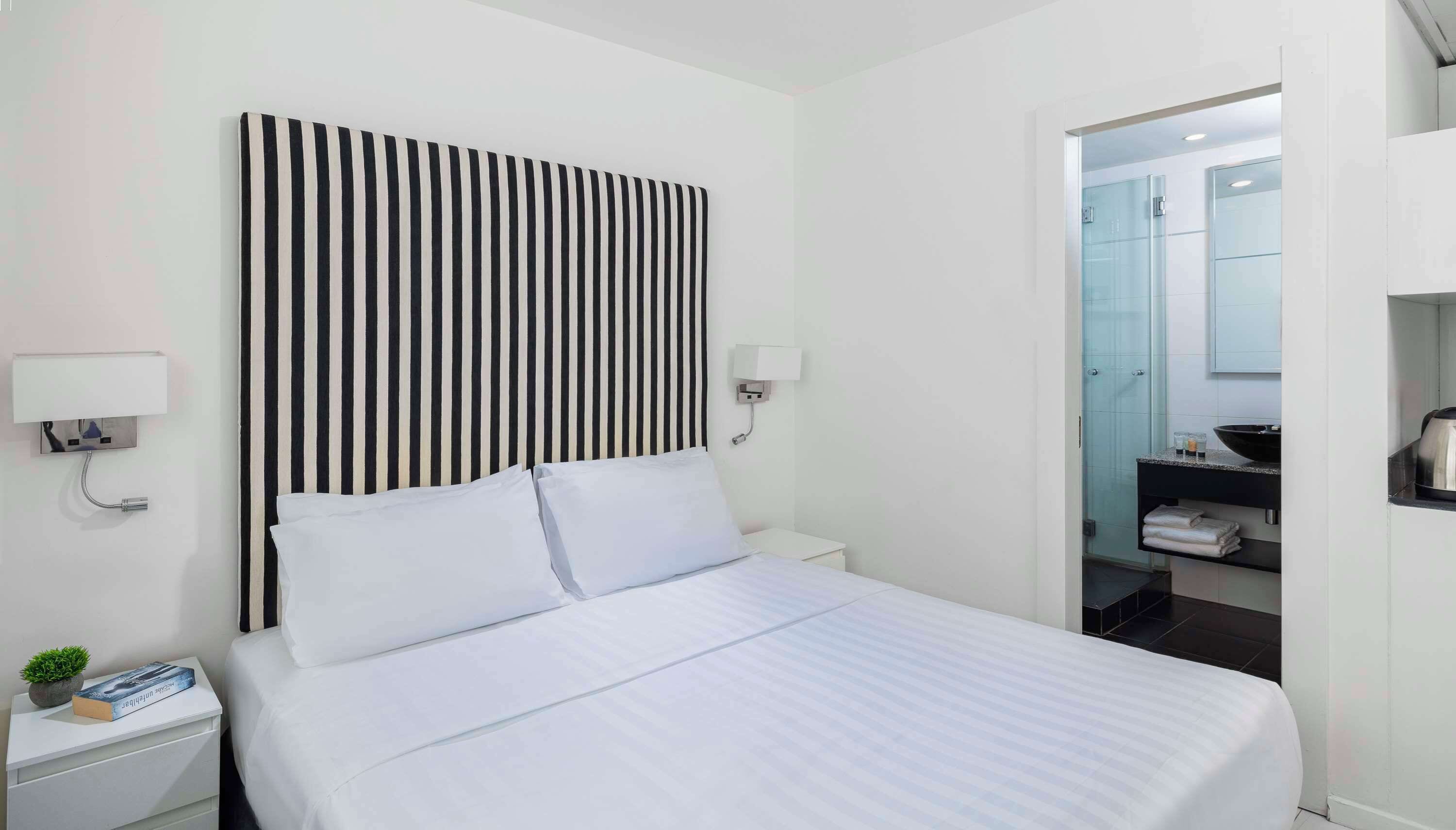 A hotel room with a large bed, a bedside table with a lamp, a wall-mounted light, and a view of the bathroom through an open door.