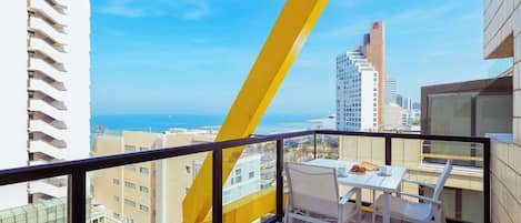 Un balcón con una mesa y sillas blancas, con vista a un paisaje urbano y al mar.