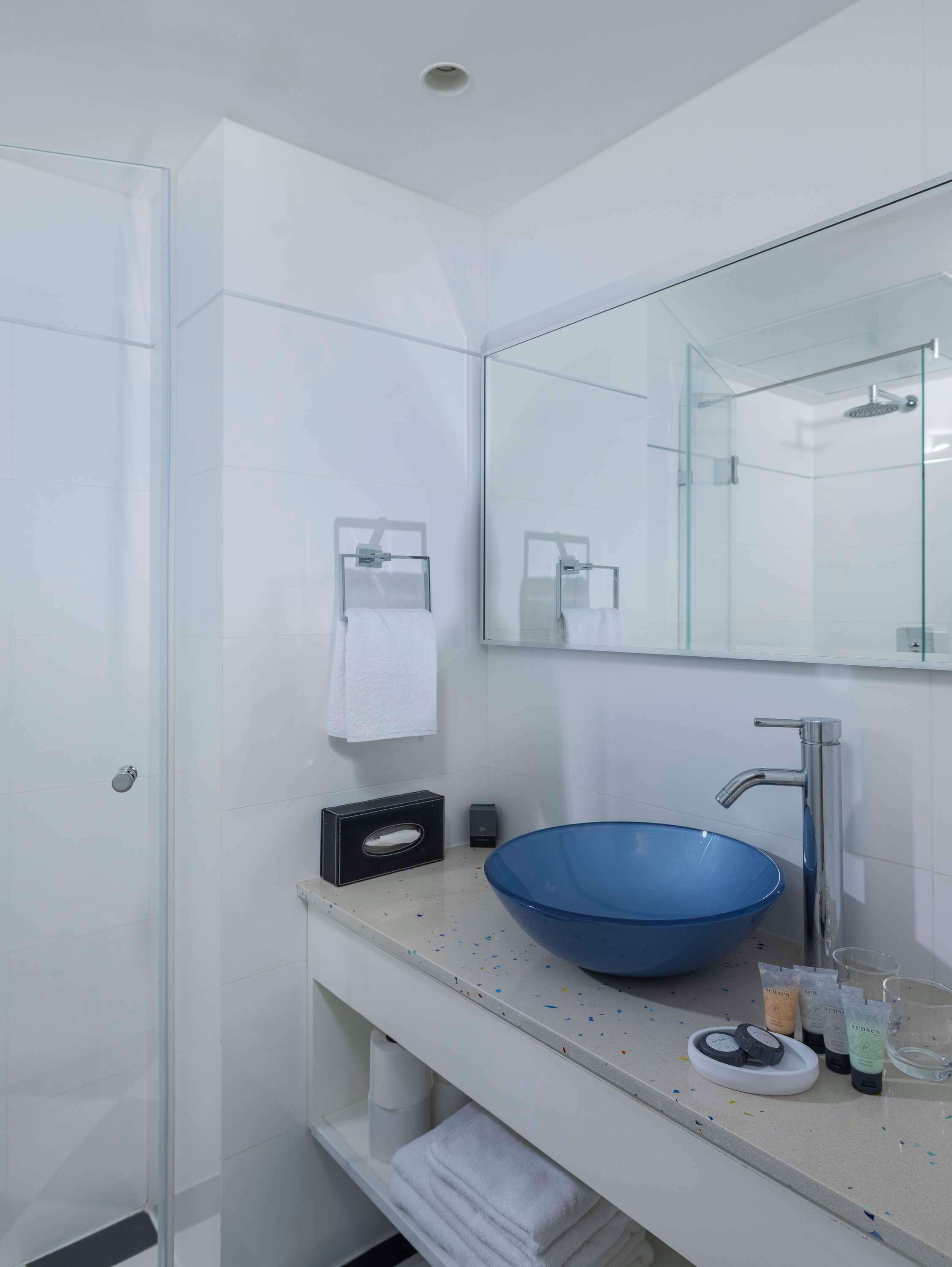 Un baño moderno con ducha envuelta en vidrio, un espejo grande y un lavabo tipo recipiente azul.