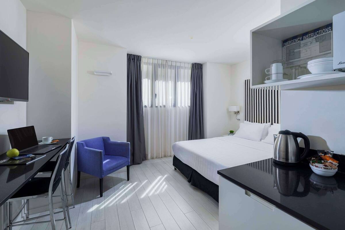 A hotel room with a bed, a desk with a laptop, a chair, a television, and a kitchenette area.