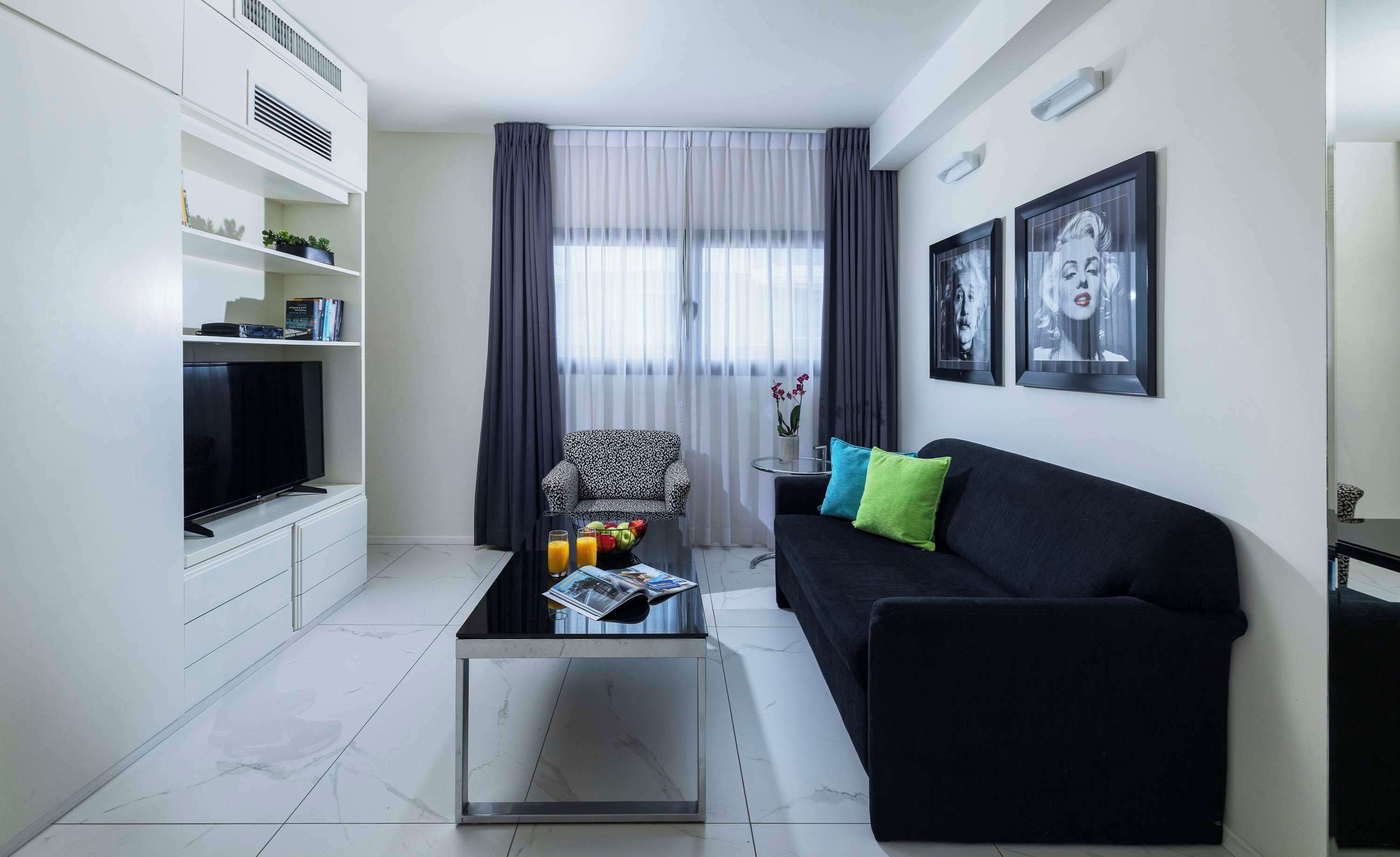 Una sala de estar moderna con un sofá negro, una mesa de centro con revistas, un televisor sobre un mueble blanco y una ventana con cortinas translúcidas.