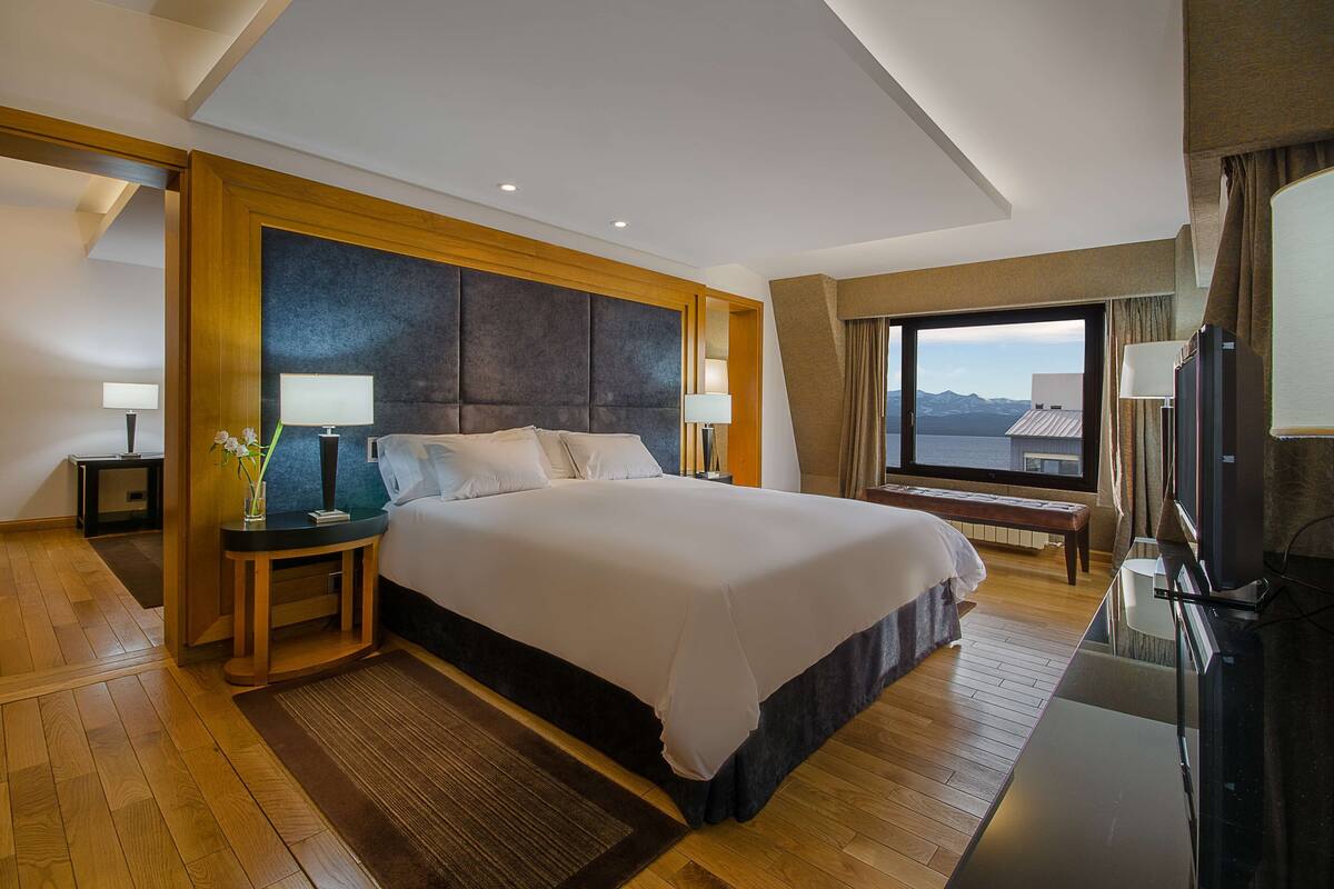 Habitación de hotel con una cama grande, cabecera de madera, dos mesitas de noche con lámparas, un televisor y vistas a las montañas por la ventana.