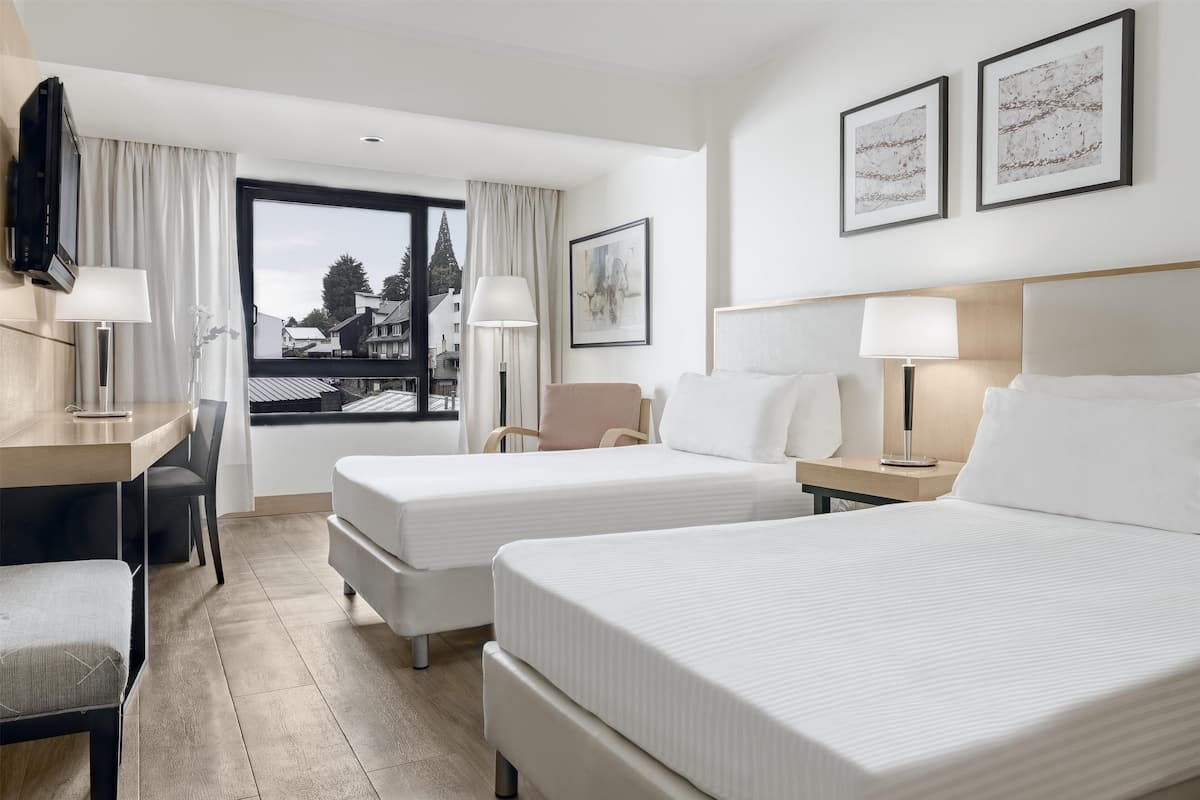 Habitación de hotel con dos camas, un escritorio, una silla y una ventana con vista a edificios.