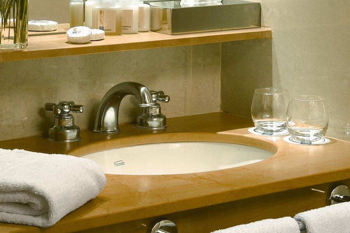 Un baño con lavamanos, dos grifos, un espejo, toallero y artículos de aseo en la encimera.