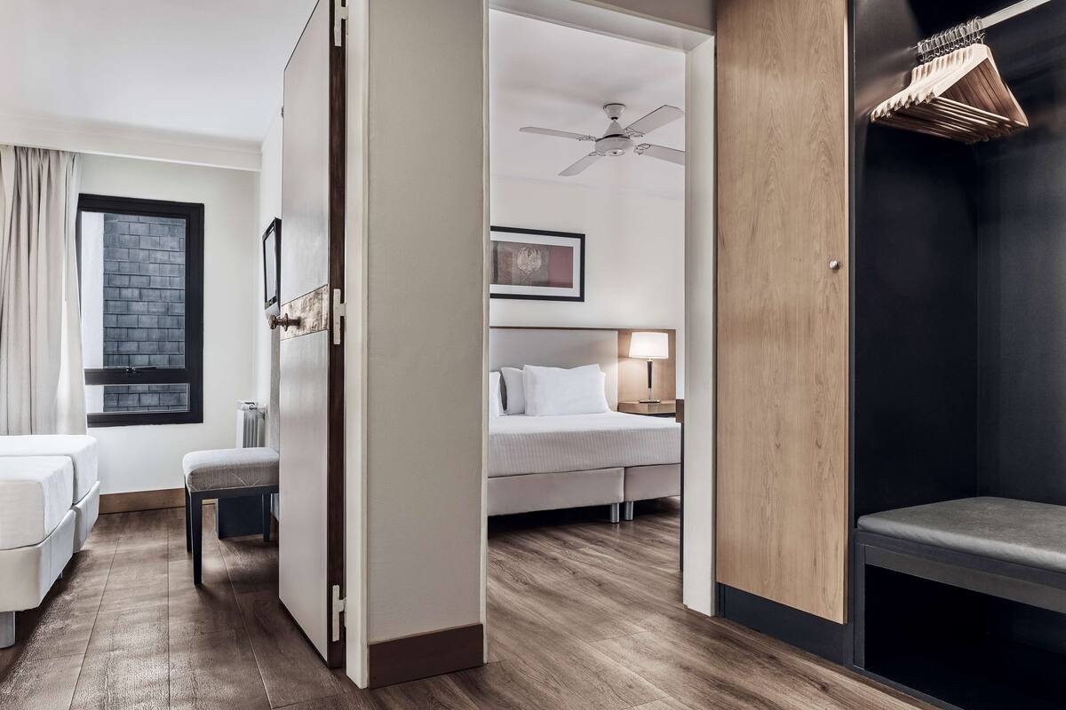 Habitación de hotel con cama, mesita de noche y una silla. Se observa una ventana con cortinas, un ventilador de techo y un panel de madera en la pared.
