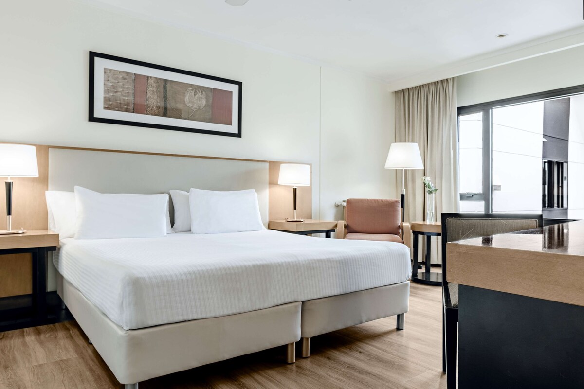 Une chambre d’hôtel avec un grand lit, deux tables de chevet, une chaise, un bureau et une grande fenêtre.