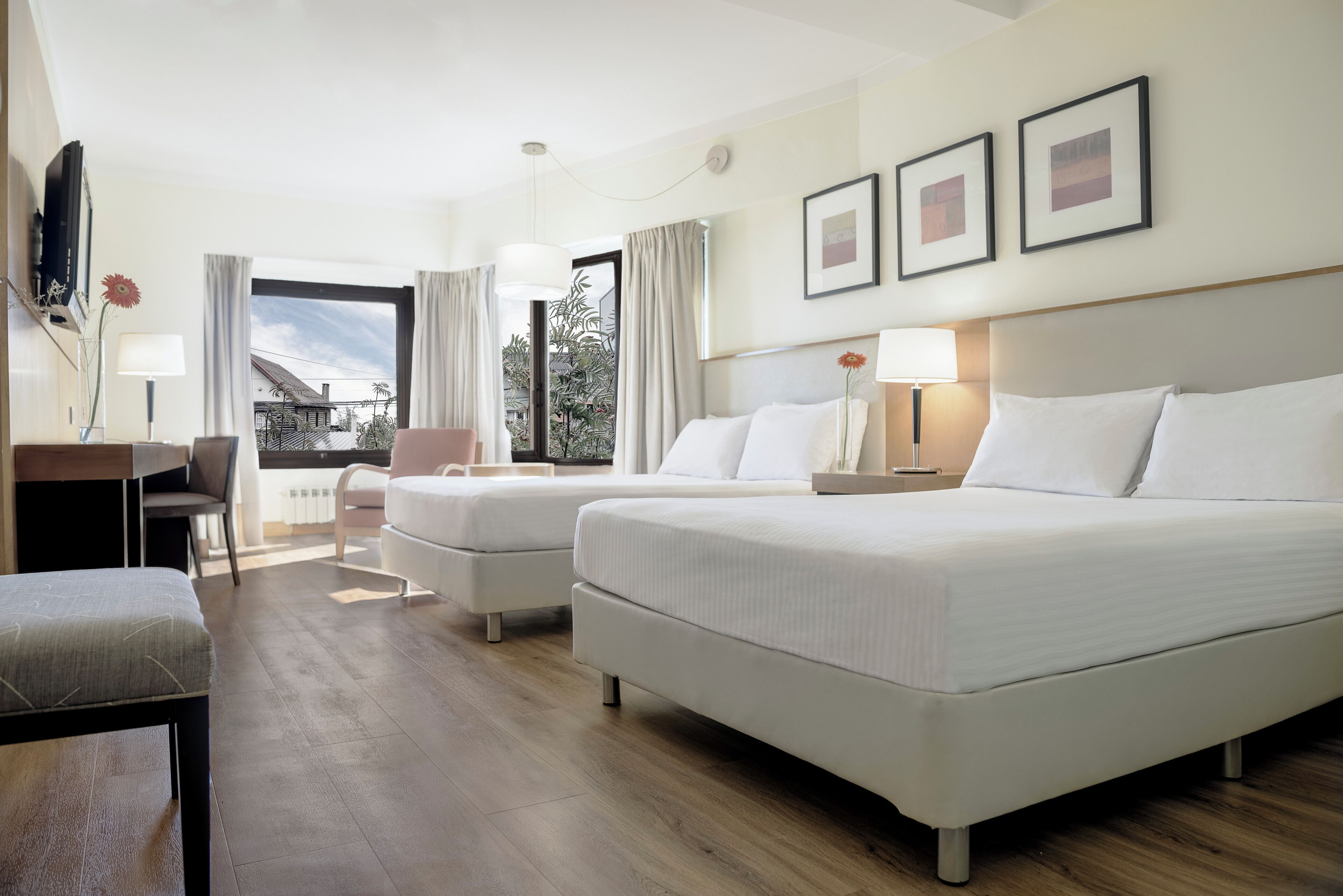 Habitación de hotel con dos camas, un escritorio, una silla, una ventana con cortinas e imágenes enmarcadas en la pared.