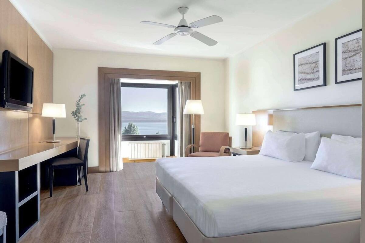 Habitación de hotel con cama, escritorio, sillas, televisor y vistas al exterior.