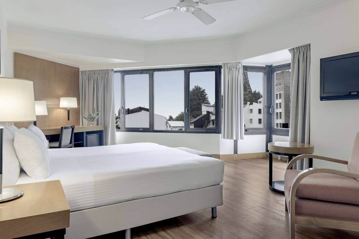 Habitación de hotel con una cama grande, un escritorio con lámpara, una silla, un televisor y vistas a edificios a través de las ventanas.