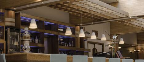 Zona de bar con estanterías de madera, lámparas colgantes y una barra con sillas.