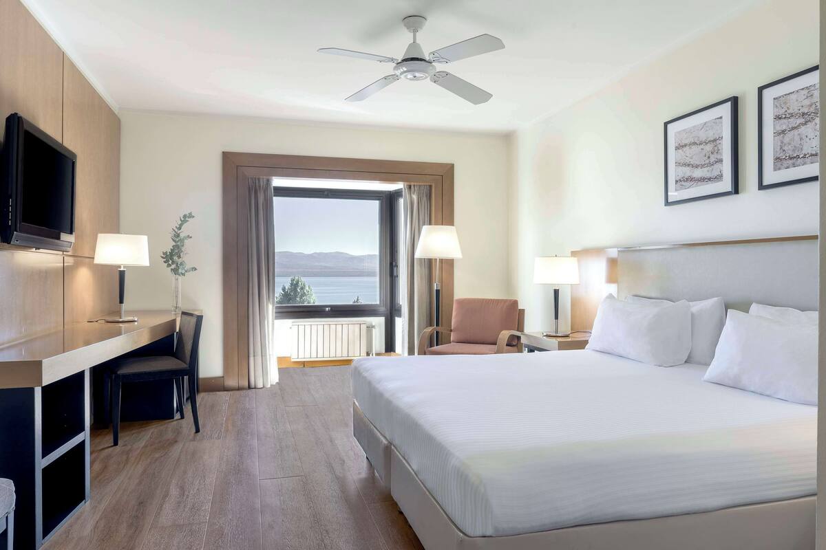 Habitación de hotel con cama, escritorio, silla, televisión y balcón con vistas.