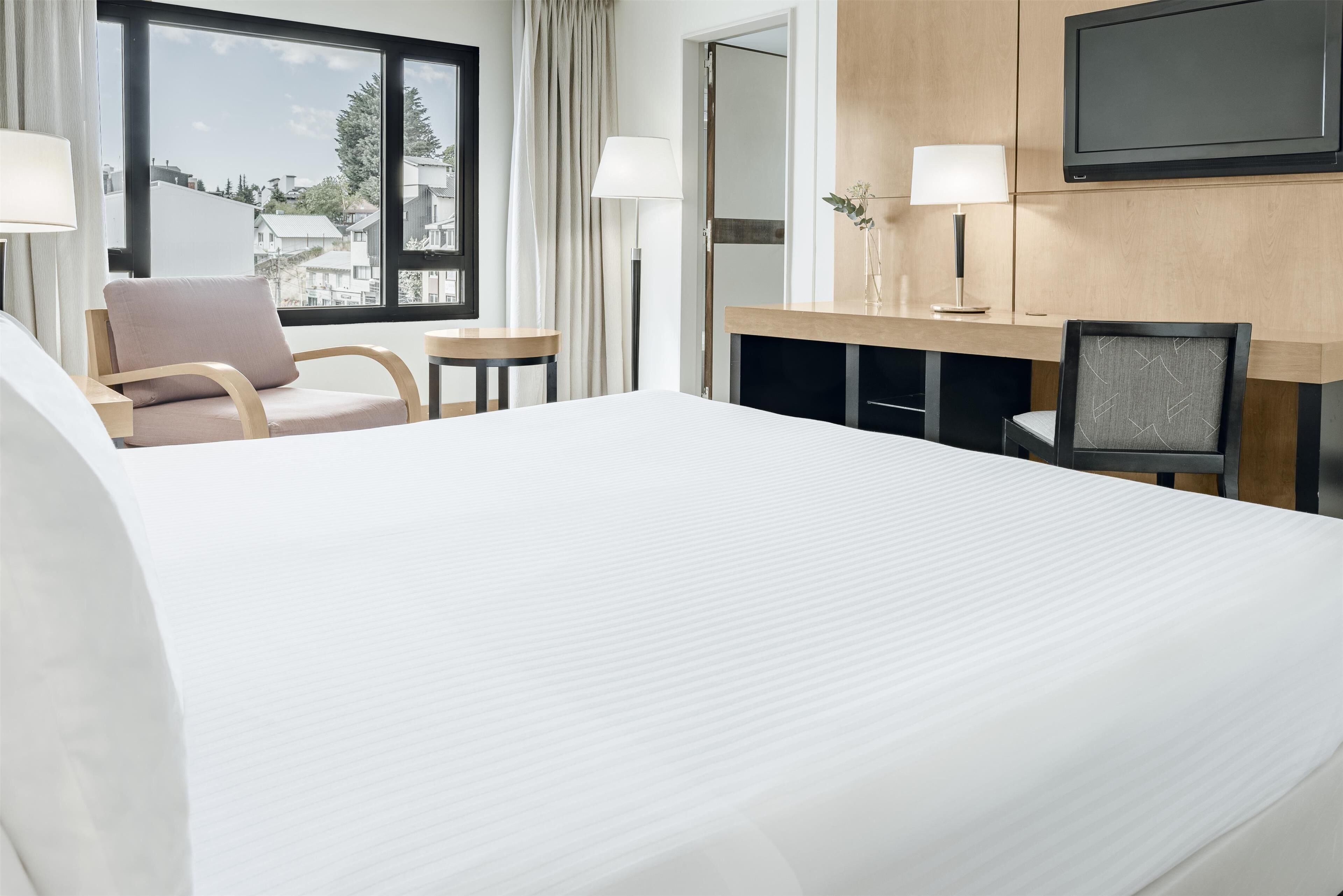 A hotel room with a large bed, a desk with a lamp, a chair, and a TV mounted on the wall.