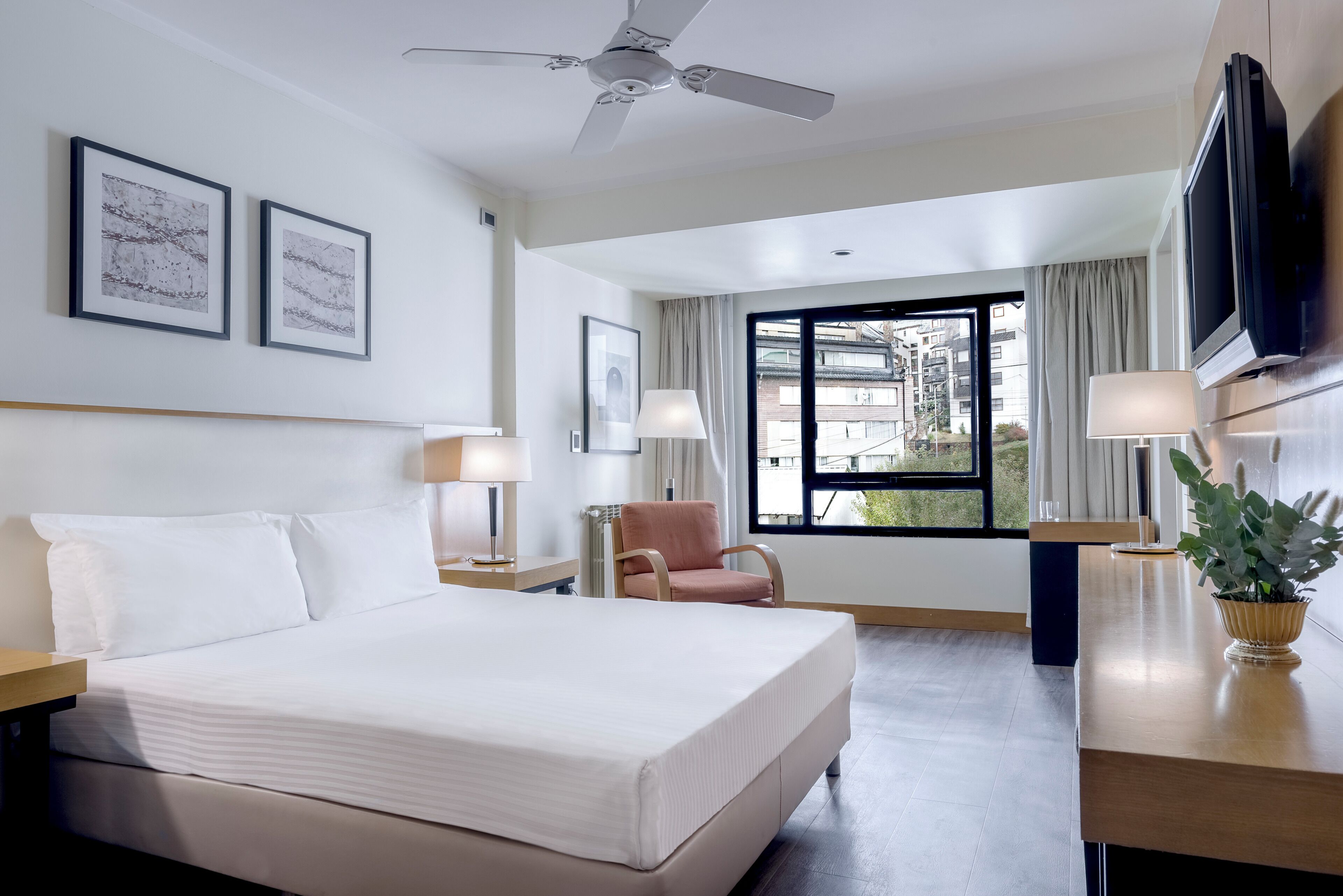 Habitación de hotel con cama, una silla, escritorio, televisión y ventana con vista a edificios.