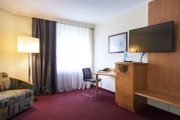 A hotel room with a sofa, a desk, a chair, a TV, and a window with curtains.