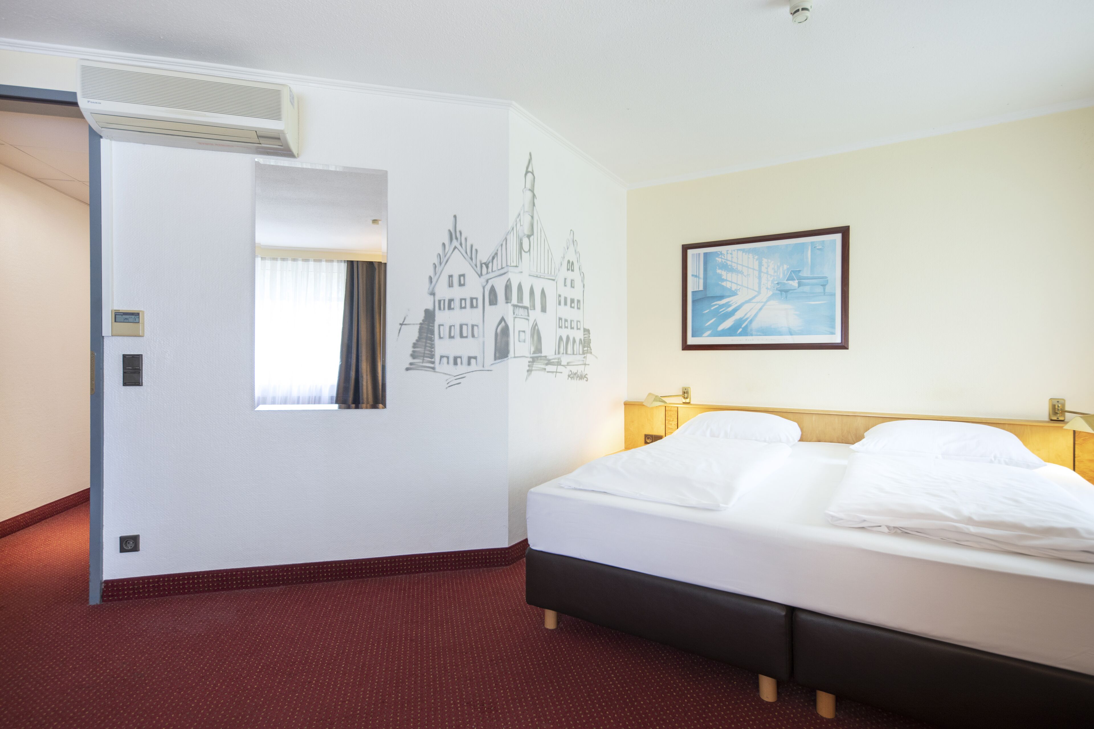 Habitación de hotel con cama, aire acondicionado de pared, ventana con cortinas, un cuadro enmarcado y un mural en la pared.