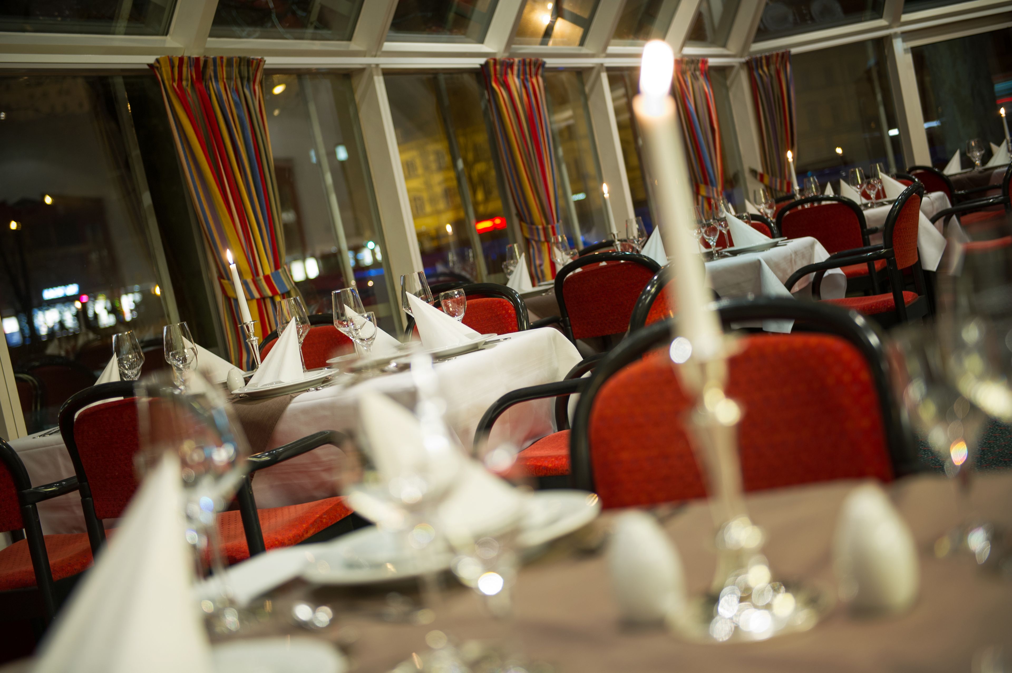 Un restaurante con mesas dispuestas para comer, sillas rojas y vista a la ciudad de noche.