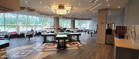A spacious indoor area with multiple tables and chairs, large windows, and a modern ceiling design.