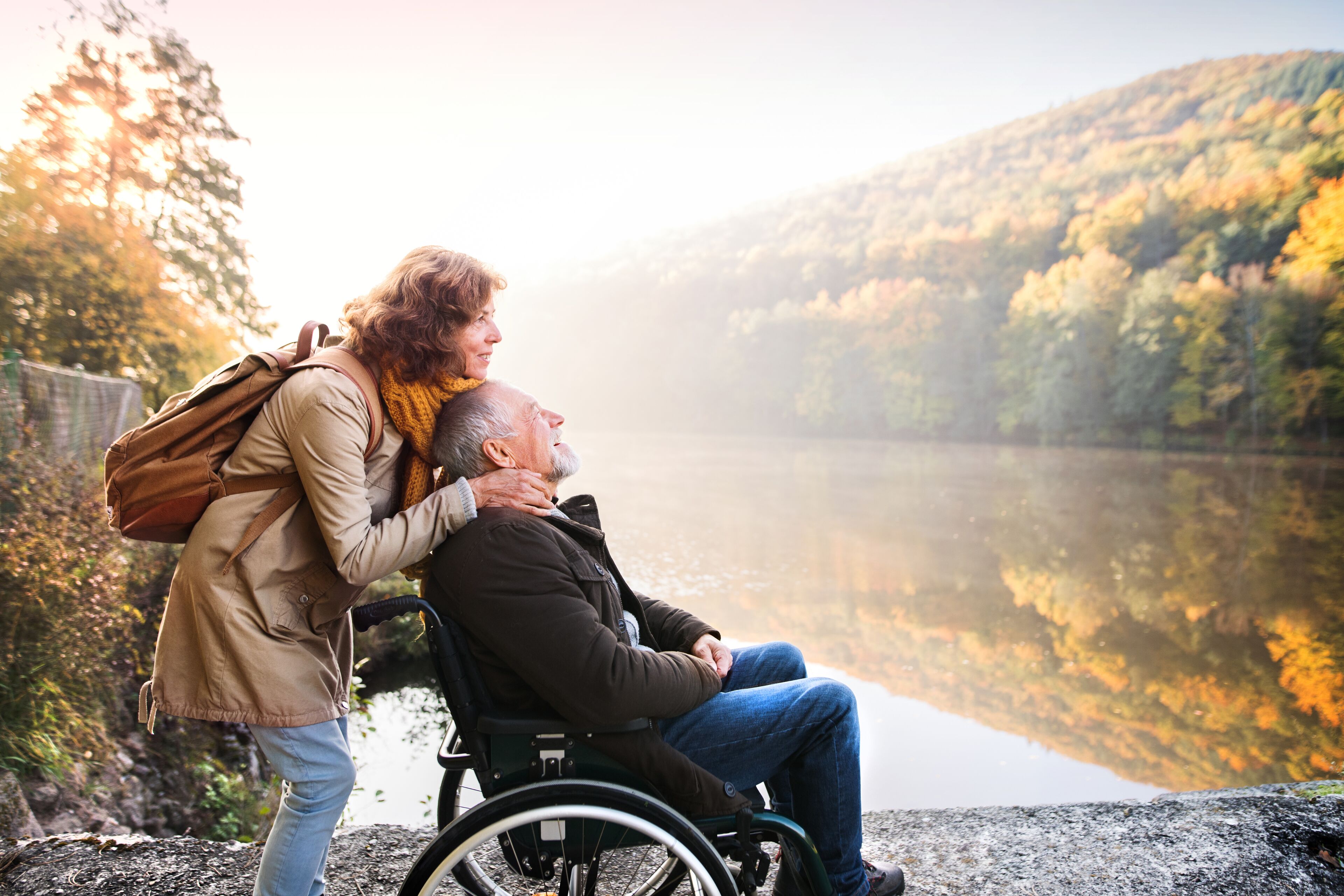 A person in a wheelchair and another person standing by a lake with a forest in the background.