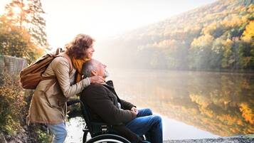A person in a wheelchair and another person standing by a lake with a forest in the background.