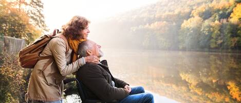 A person in a wheelchair and another person standing by a lake with a forest in the background.