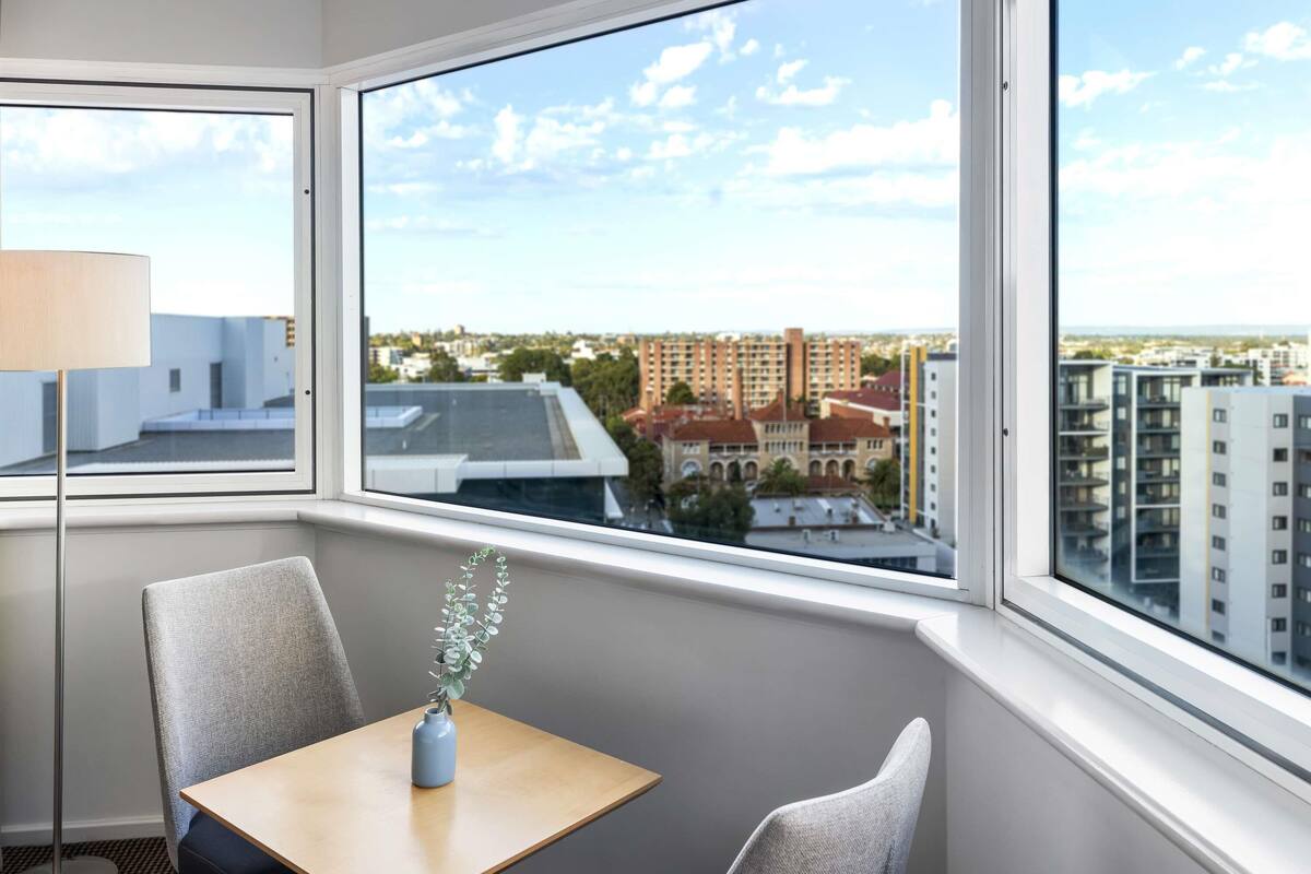 A hotel room with a large window offering a city view, a small table with a vase, and a chair.