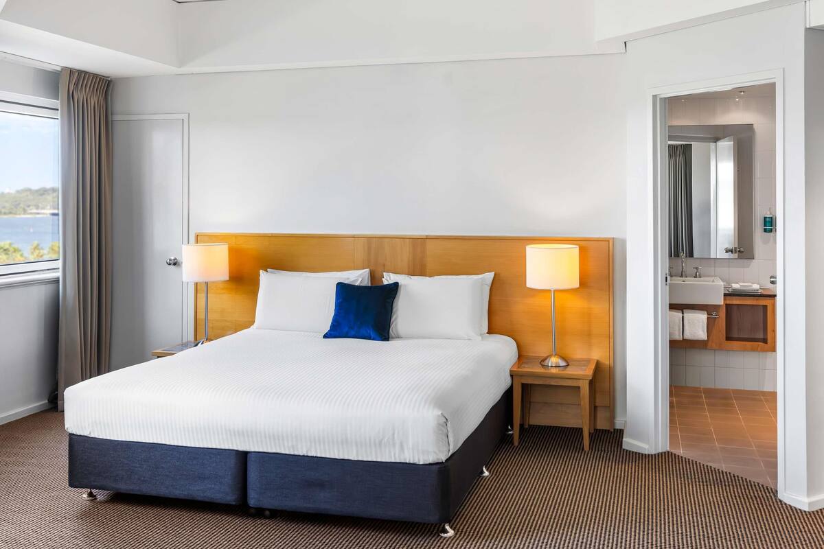 Une chambre d’hôtel avec un grand lit, deux lampes de chevet, une tête de lit en bois, une vue sur la mer, une salle de bain avec un miroir et un lavabo, et une moquette à motifs.
