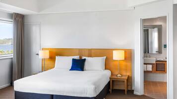 A hotel room with a large bed, two bedside lamps, a wooden headboard, a view of the sea, a bathroom with a mirror and sink, and a patterned carpet.