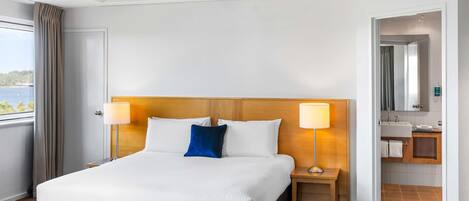 A hotel room with a large bed, two bedside lamps, a wooden headboard, a view of the sea, a bathroom with a mirror and sink, and a patterned carpet.