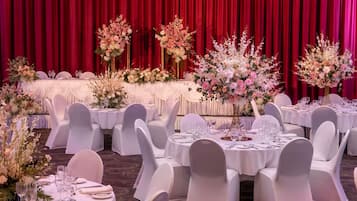 Un salón de banquetes con mesas redondas, sillas blancas y cortinas rojas.