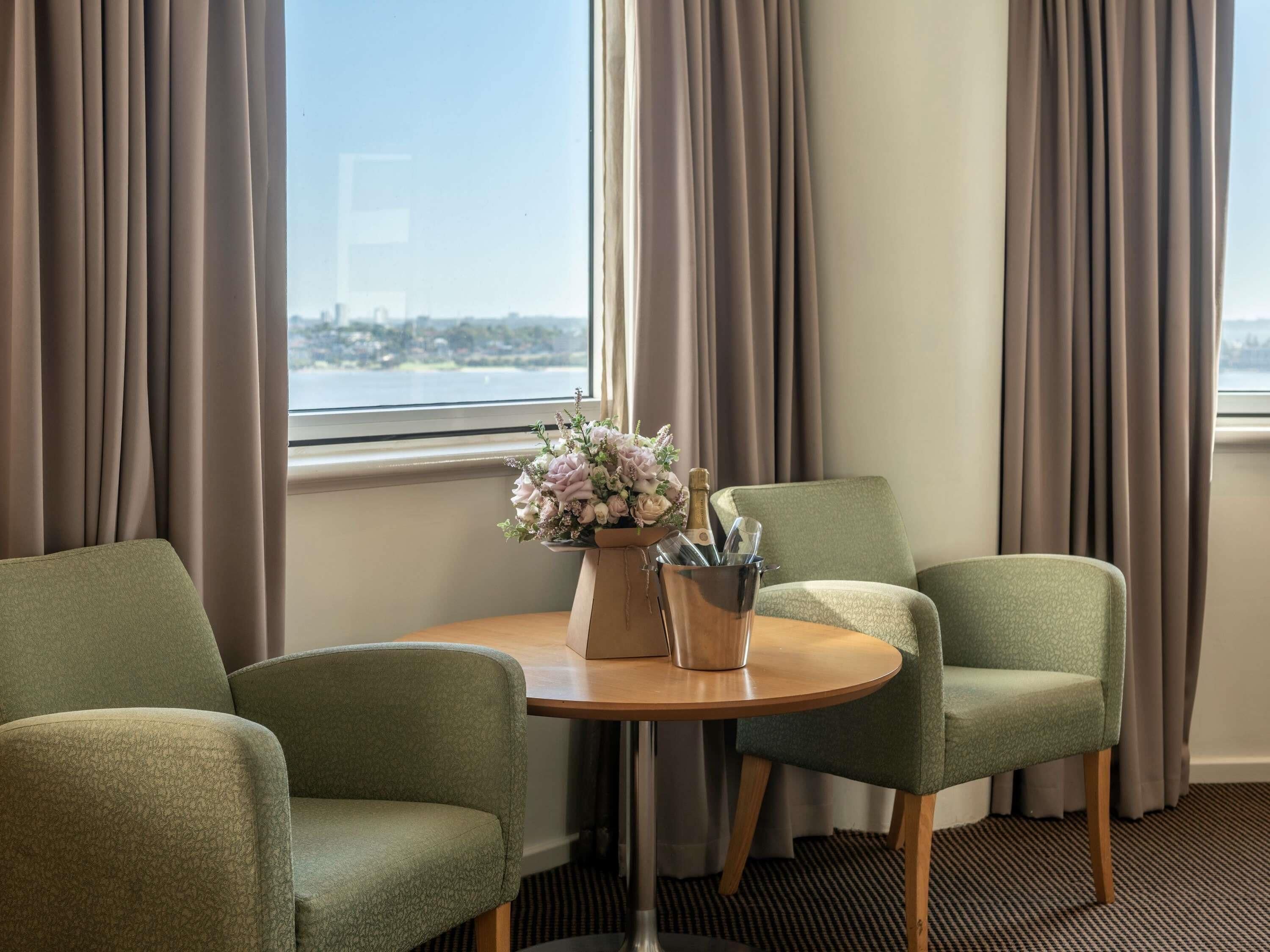 A hotel room with two green armchairs, a wooden table, a vase with flowers, and a bottle of champagne.