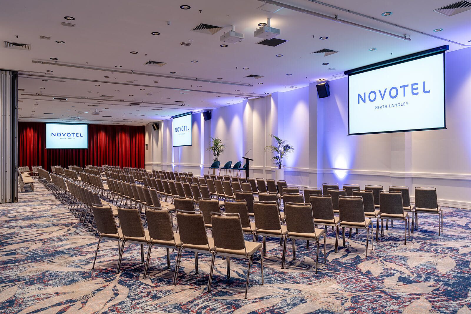 Una sala de conferencias con filas de sillas, una pantalla grande mostrando 'NOVOTEL Perth Langley' y una alfombra estampada.