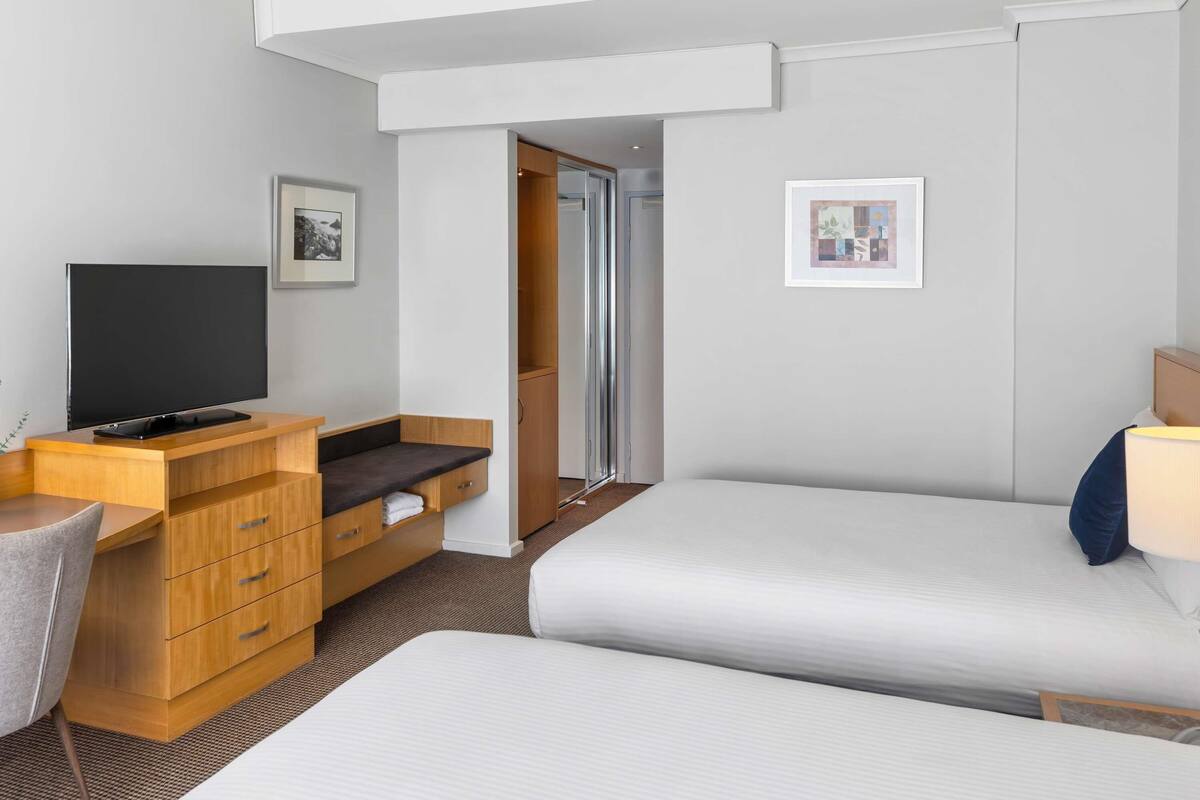 Une chambre d’hôtel avec deux lits, un bureau doté d’un téléviseur à écran plat, un meuble de rangement en bois et une armoire.