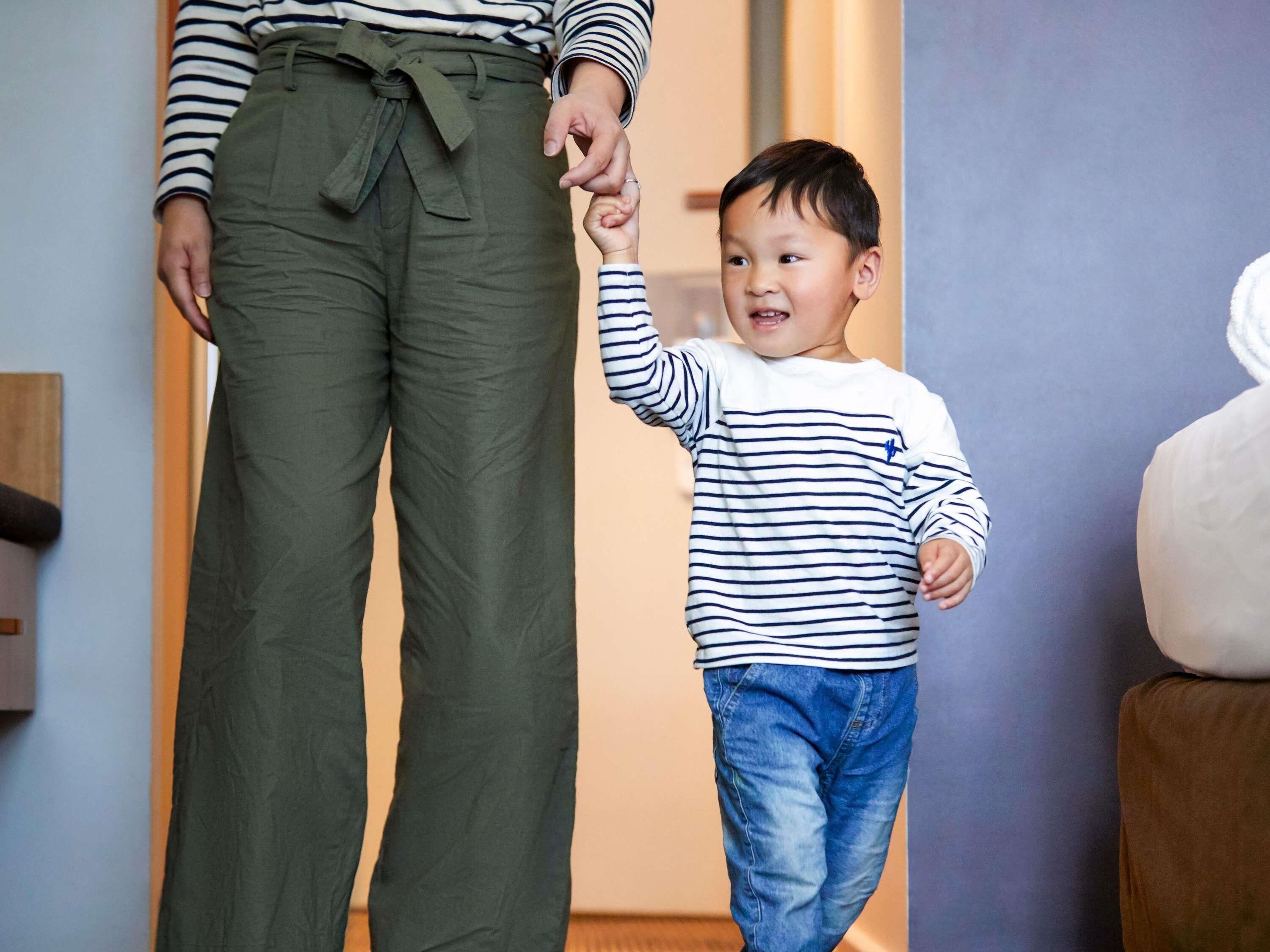 A person holding a child's hand in a room with a wooden floor and a brown couch.