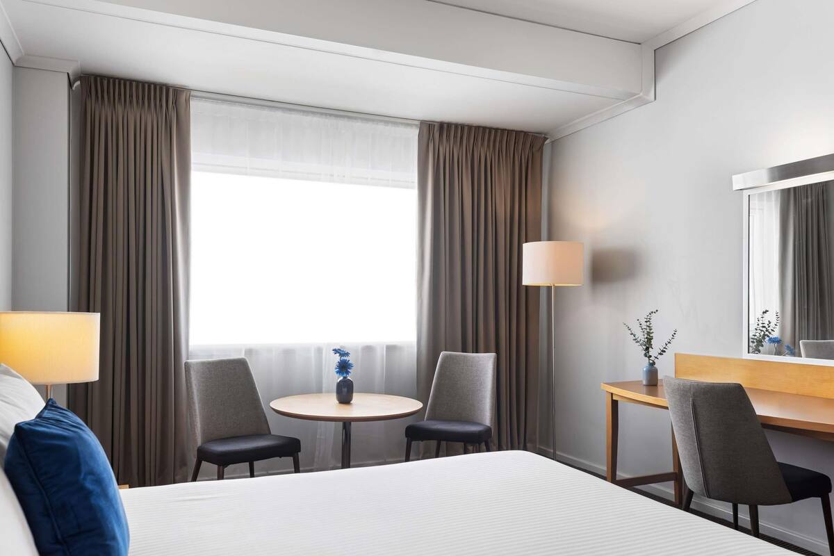 A hotel room with a large bed, two chairs, a small round table, a desk, a lamp, and a mirror.